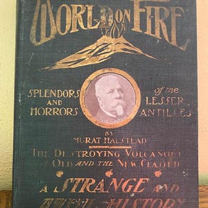 May include: Antique book titled "The World on Fire" with gold lettering on a dark blue cover. The cover features an illustration of a volcano and a portrait. The book's subtitle reads "Splendors and Horrors of the Lesser Antilles."
