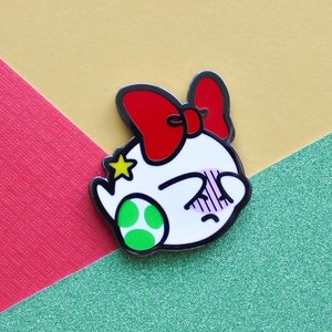 Pode incluir: Um broche de esmalte branco com um personagem de desenho animado usando um la&ccedil;o vermelho. O personagem tem uma concha verde e uma estrela na cabe&ccedil;a. O personagem est&aacute; corado e tem os olhos fechados.