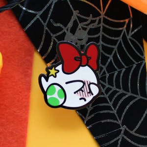 Pode incluir: Um broche de esmalte branco com um personagem de desenho animado usando um la&ccedil;o vermelho. O personagem tem um olho verde e uma express&atilde;o de surpresa. O broche est&aacute; em um fundo preto e prateado com um padr&atilde;o de teia de aranha.
