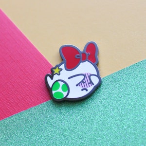 Pode incluir: Um broche de esmalte branco com um la&ccedil;o vermelho, uma estrela verde e uma concha verde. O broche apresenta um personagem de desenho animado com o rosto corado e os olhos fechados.