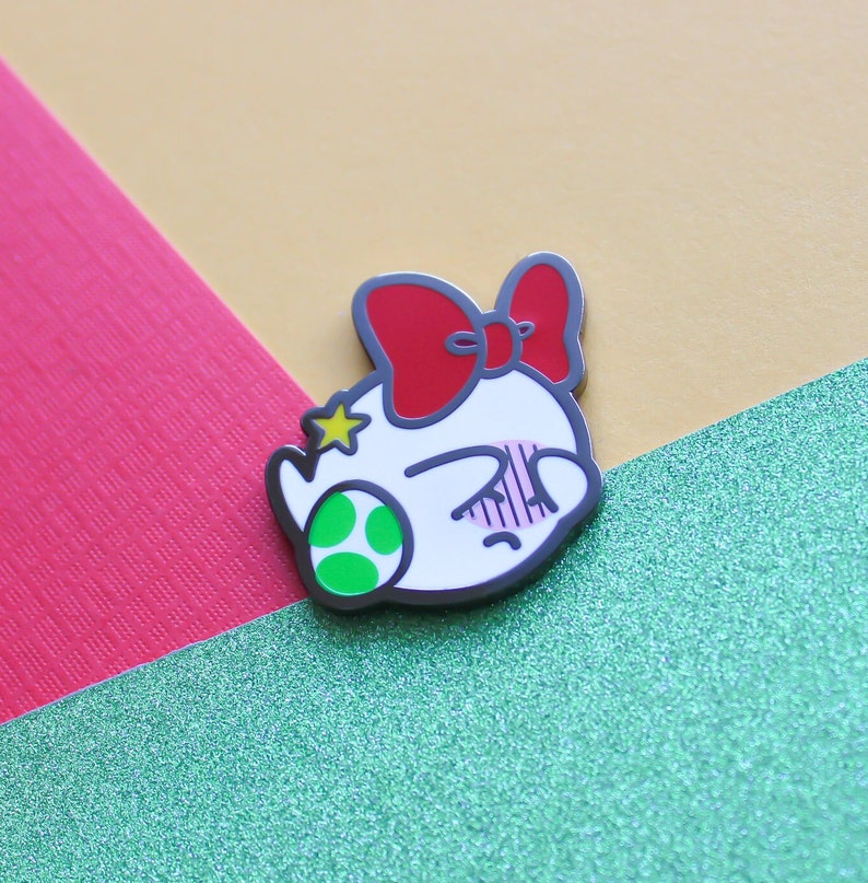 Pode incluir: Um broche de esmalte branco com um la&ccedil;o vermelho, uma estrela verde e uma concha verde. O broche apresenta um personagem de desenho animado com o rosto corado e os olhos fechados.