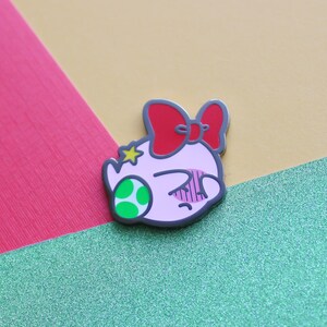 Pode incluir: Um broche de esmalte rosa e verde com um personagem de desenho animado com um la&ccedil;o vermelho e uma estrela na cabe&ccedil;a. O personagem tem uma express&atilde;o de surpresa com os olhos bem abertos e a boca ligeiramente aberta.