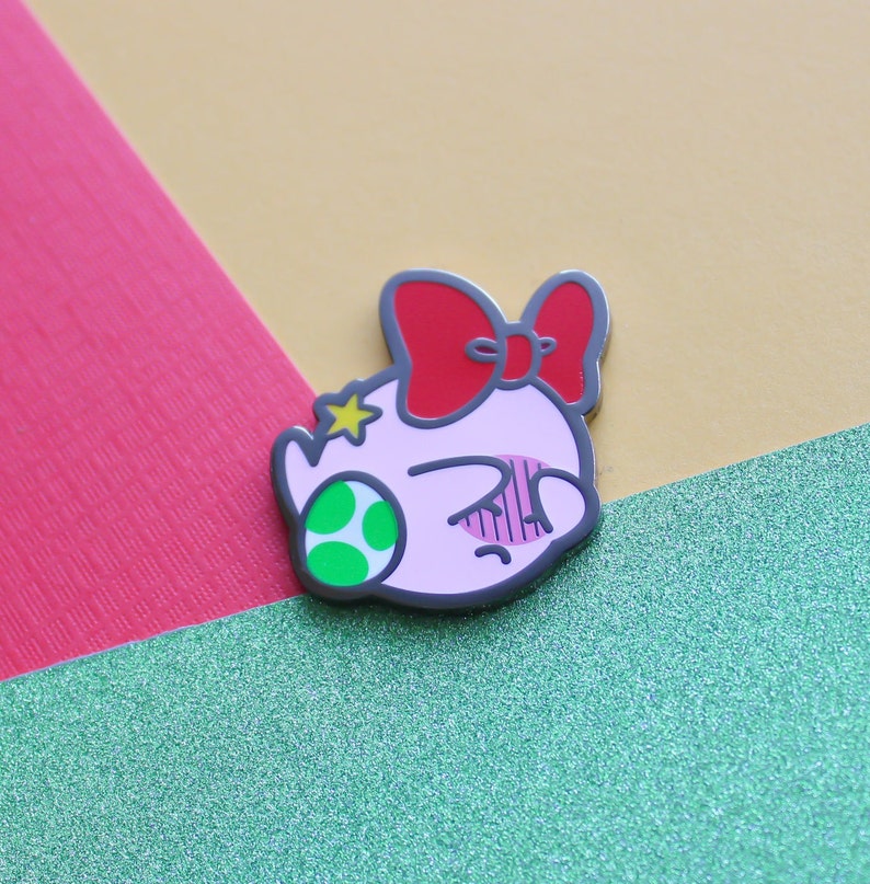 Pode incluir: Um broche de esmalte rosa e verde com um personagem de desenho animado com um la&ccedil;o vermelho e uma estrela na cabe&ccedil;a. O personagem tem uma express&atilde;o de surpresa com os olhos bem abertos e a boca ligeiramente aberta.