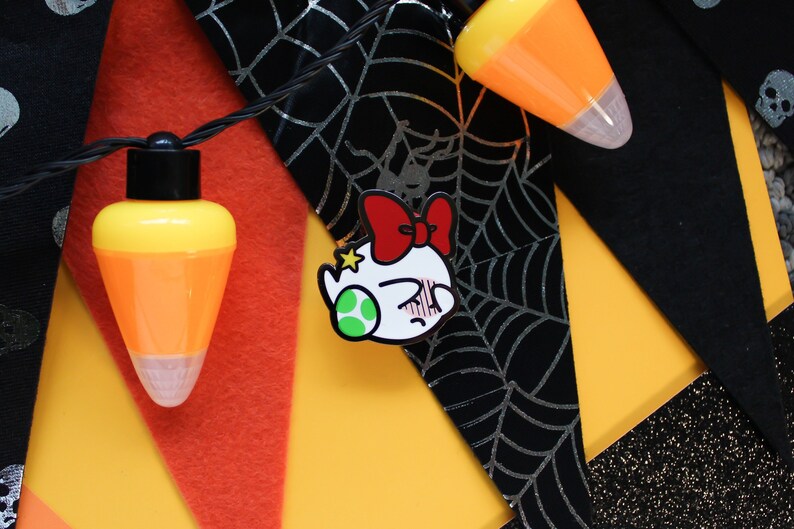 Pode incluir: Um broche de esmalte branco com um personagem de desenho animado usando um la&ccedil;o vermelho. O personagem tem um olho verde e uma express&atilde;o de surpresa. O broche est&aacute; em um fundo preto e prateado com um padr&atilde;o de teia de aranha.