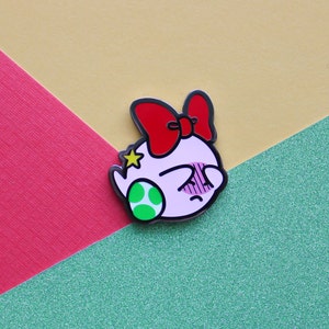 Pode incluir: Um personagem de desenho animado rosa e branco com um la&ccedil;o vermelho e uma estrela verde na cabe&ccedil;a. O personagem est&aacute; usando uma concha verde e branca. O personagem &eacute; um broche.