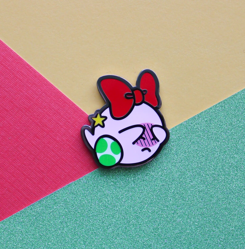Pode incluir: Um personagem de desenho animado rosa e branco com um la&ccedil;o vermelho e uma estrela verde na cabe&ccedil;a. O personagem est&aacute; usando uma concha verde e branca. O personagem &eacute; um broche.