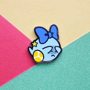 Pode incluir: Um broche de esmalte azul e amarelo com um personagem de desenho animado com um la&ccedil;o azul e uma estrela amarela. O personagem est&aacute; segurando um ovo amarelo.