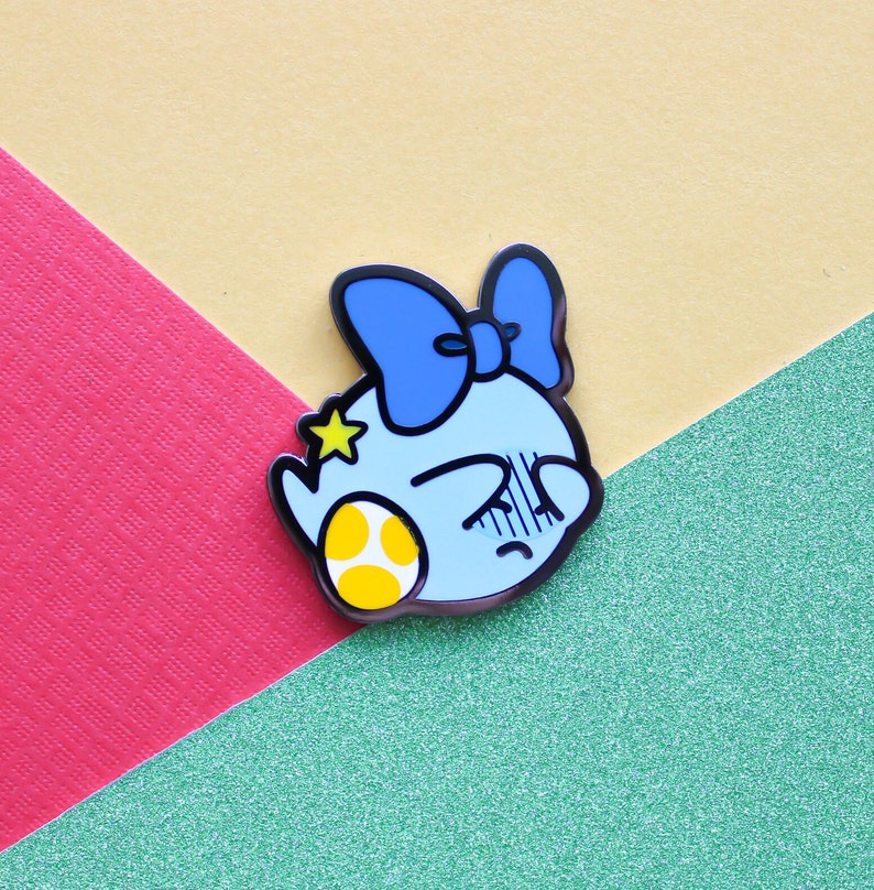 Pode incluir: Um broche de esmalte azul e amarelo com um personagem de desenho animado com um la&ccedil;o azul e uma estrela amarela. O personagem est&aacute; segurando um ovo amarelo.
