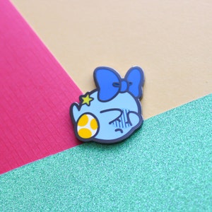 Pode incluir: Um broche de esmalte azul e amarelo com um personagem de desenho animado com um la&ccedil;o azul, uma estrela amarela e um ovo amarelo e branco com manchas. O personagem tem uma express&atilde;o de ang&uacute;stia.