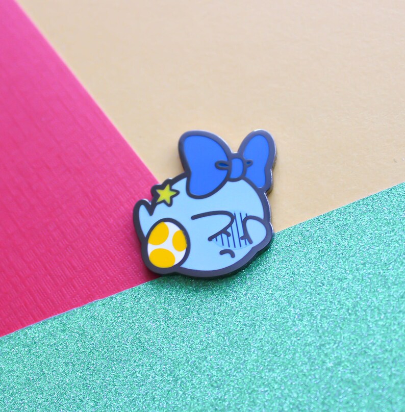 Pode incluir: Um broche de esmalte azul e amarelo com um personagem de desenho animado com um la&ccedil;o azul, uma estrela amarela e um ovo amarelo e branco com manchas. O personagem tem uma express&atilde;o de ang&uacute;stia.