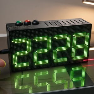 May include: A black rectangular digital scoreboard with a bright green LED display showing the number 2228. The device features three circular buttons on top, a small antenna, and is operated by a handheld remote with a wood grain finish.