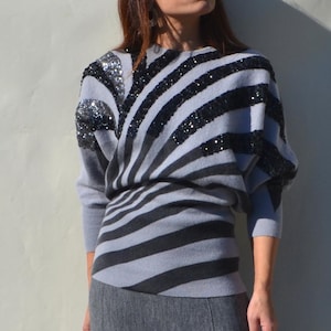 Può includere: Un maglione a righe grigie e nere con abbellimenti di paillettes sulle spalle. Il maglione è abbinato a una gonna grigia. L'outfit è un ensemble elegante, perfetto per un look alla moda.