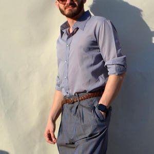 May include: A light blue button-down shirt with rolled-up sleeves, paired with gray plaid trousers and a brown leather belt. The outfit is completed with a watch. The person is wearing sunglasses.