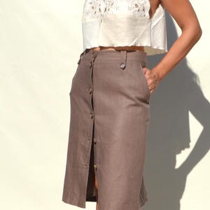 Könnte beinhalten: Ein brauner Rock mit Knöpfen, hoher Taille und Frontschlitz. Der Rock wird mit einem weißen Spitzen-Crop-Top kombiniert. Der Rock hat ein klassisches, maßgeschneidertes Design.