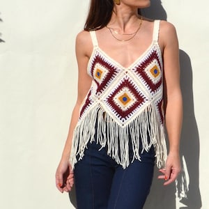 Puede incluir: Un top de crochet color crema con cuello en V y flecos. El top presenta un patrón geométrico en burdeos, amarillo y crema. Las correas son de color crema y los flecos cuelgan del dobladillo inferior.