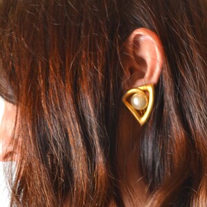 May include: A gold-toned earring with a triangular design, featuring a pearl at its center. The earring is worn on the ear of a person with long, wavy brown hair. The earring's shape is geometric and modern, creating a striking contrast.