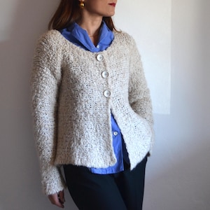 May include: A cream-coloured, textured knit cardigan with three large, round buttons. The cardigan is worn over a blue collared shirt and black trousers. The sleeves are long, and the cardigan has a slightly open front.