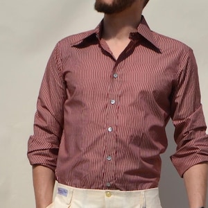 May include: A red and black striped shirt with long sleeves and a classic collar. The shirt is buttoned up and worn with cream-coloured trousers. The model is wearing a black watch. The shirt has a classic collar.