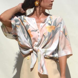 Puede incluir: Una blusa con estampado floral con la parte delantera anudada y una falda beige. La blusa presenta una paleta de colores pastel con acentos florales naranjas y amarillos. La falda es de color beige claro liso. El conjunto es de estilo vintage.