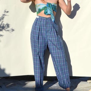 May include: Blue plaid high-waisted pants with pleats. The pants have a relaxed fit and are cuffed at the ankles. A silk scarf in shades of blue and green is worn as a top.