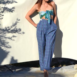 May include: A woman wearing a blue plaid high-waisted pants and a matching scarf top. The pants have a pleated front and a relaxed fit. The scarf top is tied in a bow. The outfit is set against a white backdrop.