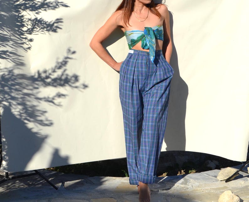 May include: A woman wearing a blue plaid high-waisted pants and a matching scarf top. The pants have a pleated front and a relaxed fit. The scarf top is tied in a bow. The outfit is set against a white backdrop.