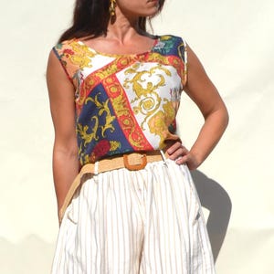 Pode incluir: Uma blusa sem mangas com um padr&atilde;o colorido em ouro, azul, vermelho e branco. A blusa &eacute; combinada com cal&ccedil;&otilde;es listrados brancos e bege e um cinto tecido. O conjunto &eacute; de estilo vintage.