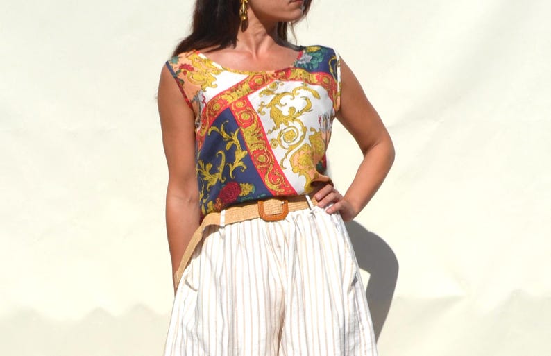 Pode incluir: Uma blusa sem mangas com um padr&atilde;o colorido em ouro, azul, vermelho e branco. A blusa &eacute; combinada com cal&ccedil;&otilde;es listrados brancos e bege e um cinto tecido. O conjunto &eacute; de estilo vintage.