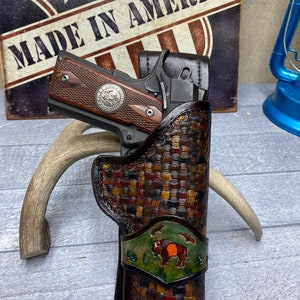 May include: A brown leather holster with a woven pattern and a buffalo design. The holster is holding a silver and black handgun with a wooden grip. The holster has the text "Made in America" on it.