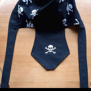 Pode incluir: Cobertura de cabeça preta com um design de caveira e ossos cruzados. A cobertura de cabeça tem um patch de caveira e ossos cruzados na frente e um padrão repetido de caveira e ossos cruzados na parte superior.