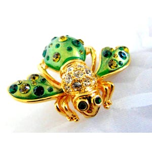 Peut inclure: Broche dorée en forme d'abeille, avec des ailes et un corps en émail vert. Le corps de l'abeille est incrusté de cristaux transparents, et les ailes et le corps présentent des accents verts et jaunes. La broche a une fermeture à épingle.
