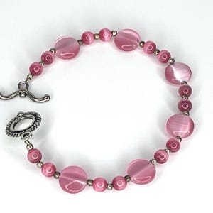 Könnte beinhalten: Ein rosa Perlenarmband mit Knebelverschluss. Das Armband hat eine Mischung aus runden und ovalen Perlen in verschiedenen Größen, die mit kleinen silberfarbenen Perlen aufgereiht sind. Der Knebelverschluss ist silberfarben.