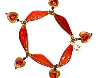 May include: A red and gold bracelet with heart-shaped charms. The bracelet features elongated red beads connected by gold-toned metal links. Each end has a gold heart charm with a red center. A tag with the text "Lalo Orna" is attached.