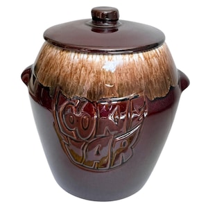 Puede incluir: Un tarro de galletas de cerámica marrón con tapa. El tarro tiene un acabado brillante y un diseño de dos tonos, con un esmalte marrón claro en la parte superior y un esmalte marrón oscuro en la parte inferior. La palabra "Cookie Jar" está grabada en relieve en la parte delantera.