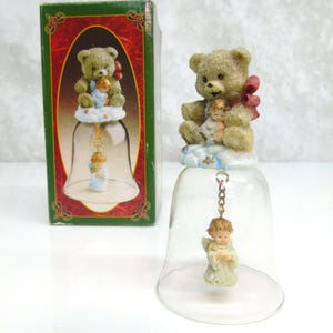 May include: A decorative bell featuring a teddy bear with a red bow, angel figurines, and a cloud design. The bell is made of clear glass and is suspended from a gold-colored chain. The bell is packaged in a green box with a red border.