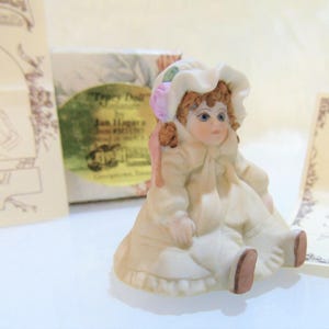 Könnte beinhalten: Miniatur-Porzellanpuppe, sitzend, mit cremefarbenem Kleid und Häubchen. Die Puppe hat rotes Haar, blaue Augen und ein rosa Band. Eine Schachtel und ein Zertifikat befinden sich im Hintergrund.
