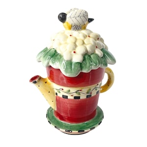 May include: A decorative ceramic teapot with a red base, yellow spout, and a lid shaped like a flower with a small bird on top. The teapot has a green and white floral design around the middle and sits on a green saucer.