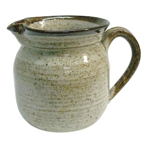 May include: A speckled ceramic pitcher with a rounded body, a curved handle, and a spout. The pitcher is primarily beige with brown accents on the rim and handle. The handmade pottery has a rustic, textured appearance.