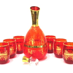 May include: A red glass decanter set with gold rims and accents. The set includes a decanter, a stopper, and six matching glasses. The glasses and decanter feature a gold floral design.