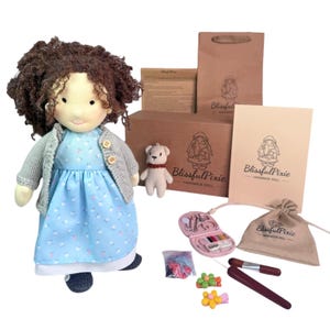 May include: A handmade doll with curly brown hair, wearing a light blue floral dress and a gray knit cardigan. The set includes a small knit bear, a brown cardboard box, a paper bag, a booklet, a drawstring pouch, and sewing accessories.