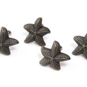 May include: Four bronze-colored starfish cabinet knobs. Each knob has a detailed, textured surface and a small silver-colored screw post. The knobs are arranged against a white background, ideal for coastal or nautical decor.