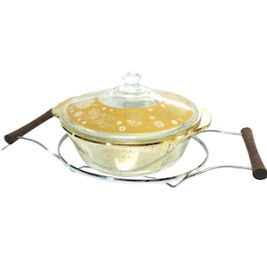 Op de afbeelding: Een vintage glazen ovenschaal met een doorzichtig deksel met een gouden bloemenontwerp. De schaal staat in een zilveren metalen standaard met houten handvatten. Ontworpen om te serveren en te koken.