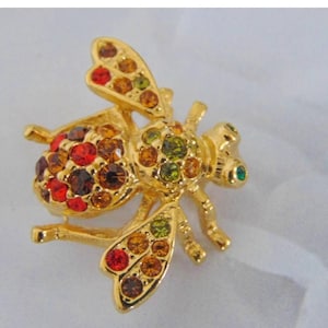 May include: A gold-coloured bee brooch adorned with colourful rhinestones. The brooch features red, orange, brown, and green stones on the body and wings. The bee has a small green rhinestone eye and detailed legs and antennae.