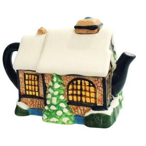 May include: A ceramic teapot shaped like a snow-covered cottage. The teapot features a black handle and spout, with windows that appear to be lit from within. A small green Christmas tree is visible on the side.