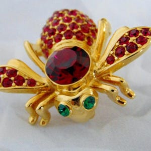May include: A gold-toned bee brooch featuring a large, round, faceted red crystal on the thorax, a body covered in small red rhinestones, and wings accented with red stones. The head has two small, round, green crystal eyes.