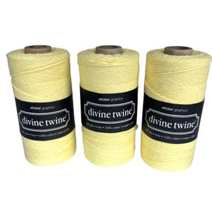 May include: Three spools of pale yellow "Divine Twine" are displayed. Each spool has a black label with white text that reads "whisker graphics" and "divine twine". The twine is 100% cotton and made in the USA.