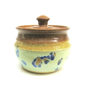 May include: A handcrafted ceramic jar with a lid. The jar features a light green base with blue and brown accents. The middle section is light yellow, and the top and lid are brown. A decorative kitchen item.