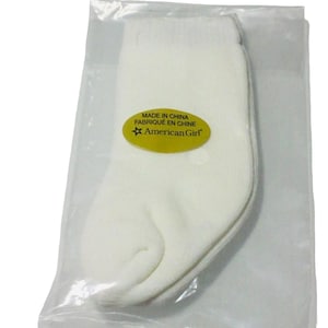 May include: A pair of plain white socks enclosed in a clear plastic bag. An oval yellow sticker is attached to the front of the packaging, featuring the text MADE IN CHINA, FABRIQUÉ EN CHINE, and the American Girl brand logo with a star symbol.
