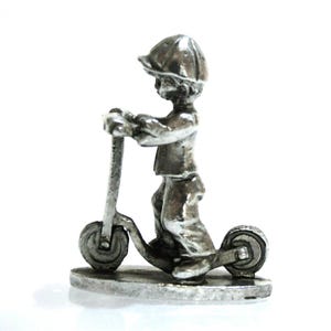 May include: A small, silver-colored figurine of a child riding a scooter. The child wears a cap and is holding onto the scooter's handlebars. The scooter has two wheels and is mounted on a small, round base.