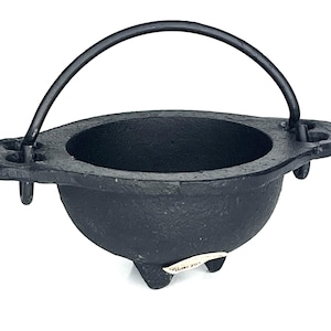 May include: A black cast iron cauldron with a rounded bowl, three short legs, and a curved metal handle attached to two side lugs. The exterior surface has a textured finish, and a small white label is visible on one of the legs.